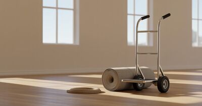A hand truck and a rolled moving blanket in the corner of an empty living room with tall windows